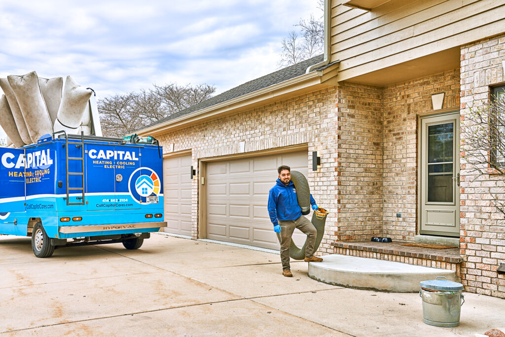 A technician walks toward the house carrying a hose for HVAC service. A blue Capital Heating & Cooling truck is parked nearby, ready to ensure your home stays comfortable with expert heating and cooling solutions.