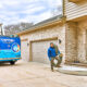 A technician walks toward the house carrying a hose for HVAC service. A blue Capital Heating & Cooling truck is parked nearby, ready to ensure your home stays comfortable with expert heating and cooling solutions.