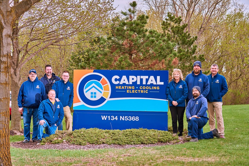 People in blue jackets pose around a Capital Heating, Cooling, and Electric sign in a grassy area with trees in the background, showcasing their expertise in maintaining year-round comfort.