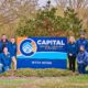 People in blue jackets pose around a Capital Heating, Cooling, and Electric sign in a grassy area with trees in the background, showcasing their expertise in maintaining year-round comfort.