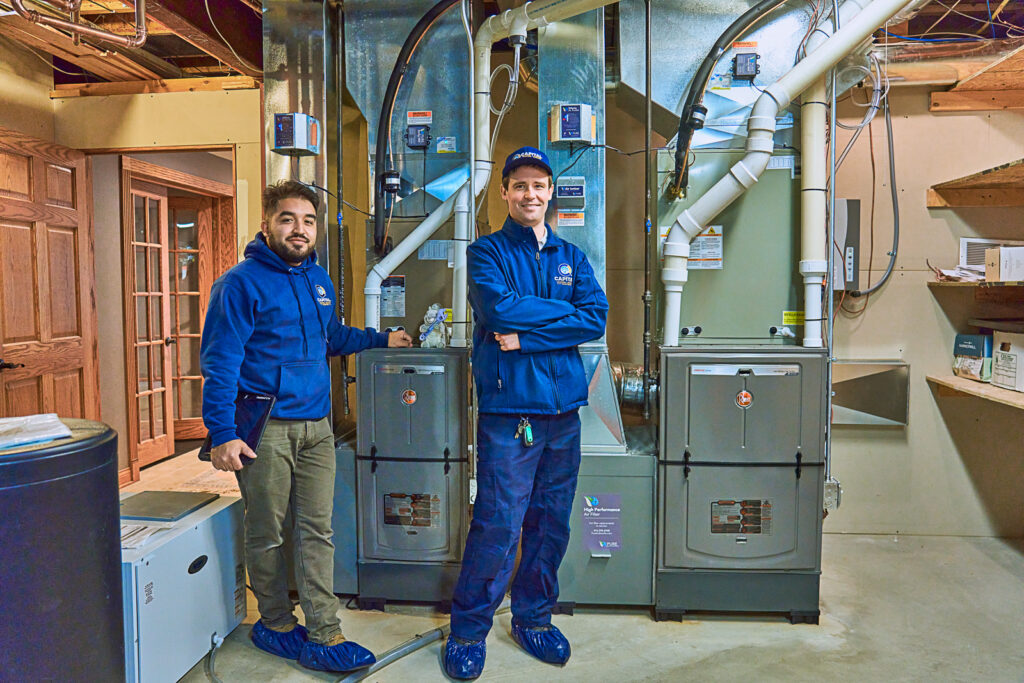 Wearing blue uniforms, two technicians stand in front of large HVAC systems in a basement. One technician points at the equipment while they discuss the complex electric components that control both heating and cooling functions.
