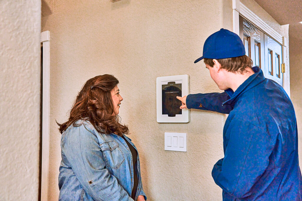 A person in a blue uniform demonstrates the functions of an electric wall-mounted device to a woman by showing her the screen.