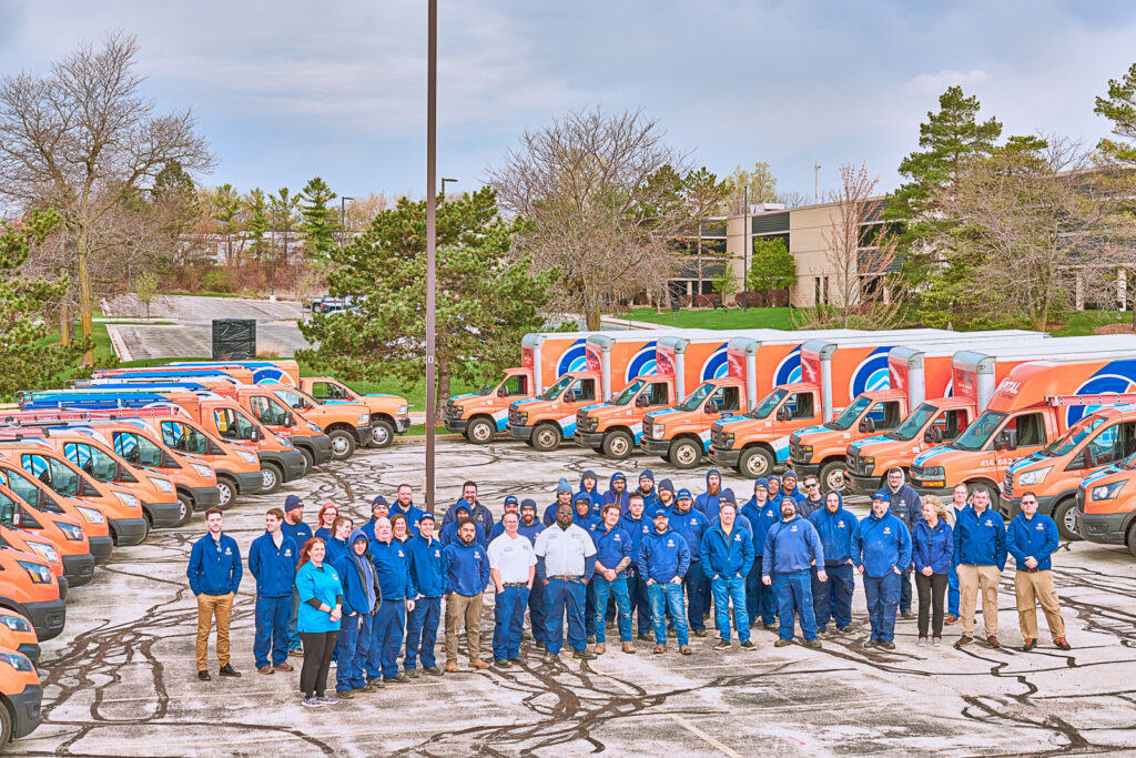 People in blue uniforms stand in a parking lot surrounded by orange and blue heating and cooling vans. Trees and a building frame this typical scene from Milwaukee.