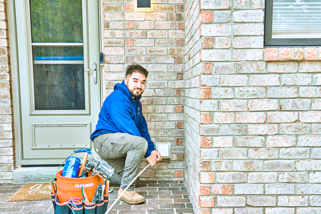 A person in a blue hoodie and work boots kneels by a brick wall and holds electric tools. A tool bag sits nearby on the brick pathway.